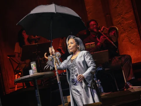 A performer in a shiny silver outfit holds an open black umbrella on stage, with other musicians in the background.