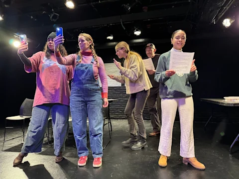 Five people are on a stage; two are holding up phones, one holds a script and faces forward, another reads a script while smiling, and one stands behind gesturing with their hands.