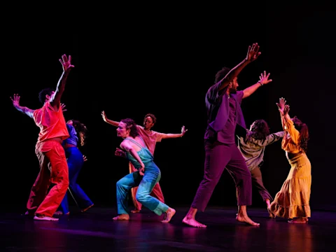 A group of dancers in colorful outfits perform on a dark stage with dramatic, expressive arm movements.