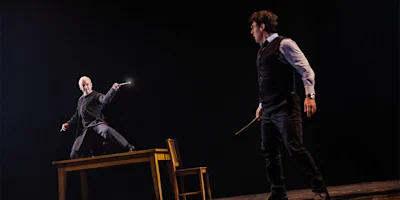 Two people holding wands face each other on a dark stage; one person stands on a table in a dramatic pose, while the other stands on the floor.