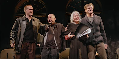 Four people stand on stage in front of arched windows, smiling and holding hands while the audience applauds.