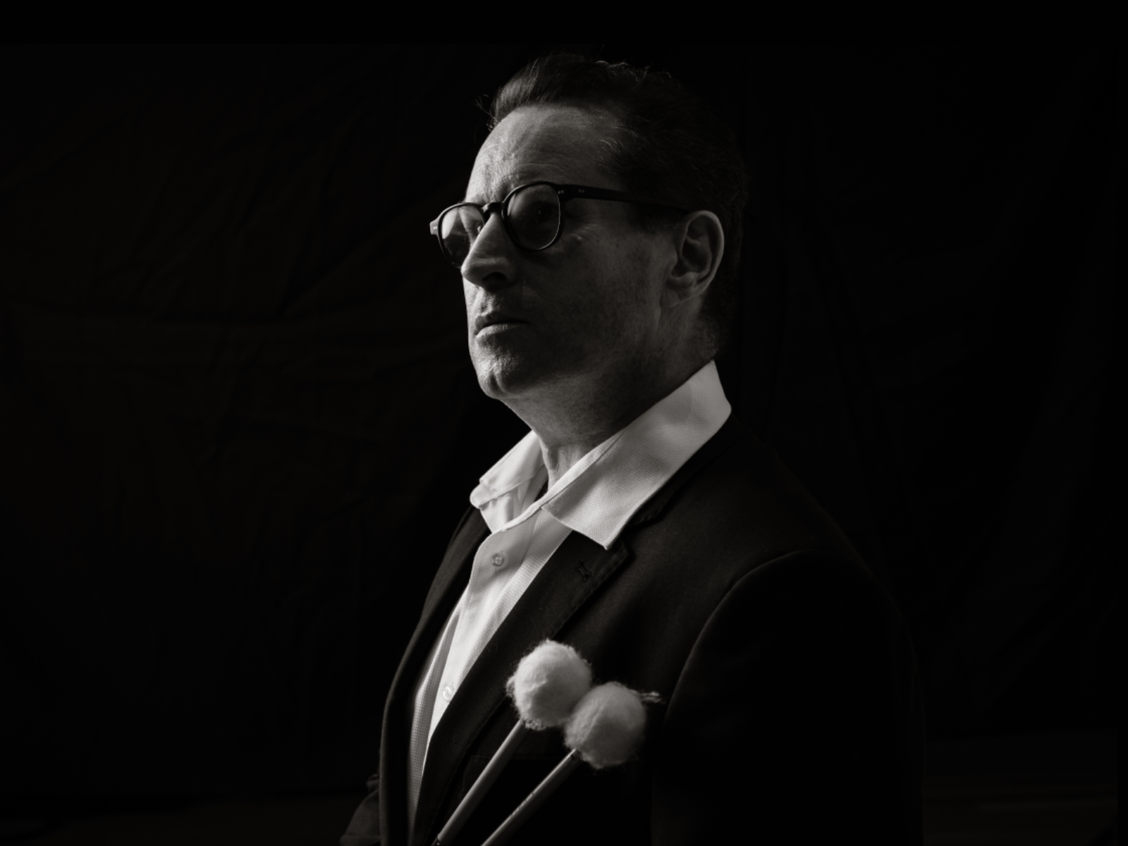 A man in glasses and a suit holds two mallets, staring forward in a black and white portrait with a dark background.