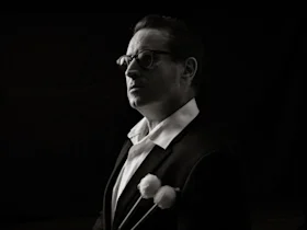 A man in glasses and a suit holds two mallets, staring forward in a black and white portrait with a dark background.