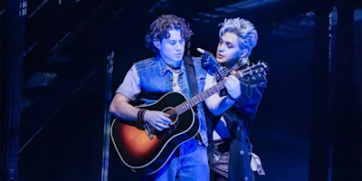 Two young men stand close together on stage, one playing an acoustic guitar while the other gestures toward him, both under blue lighting.