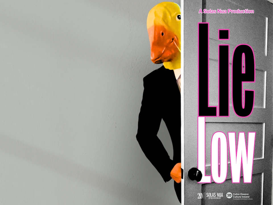 A person in a suit and yellow duck mask stands behind a door. Large pink and white text on the right reads “Lie Low.”.