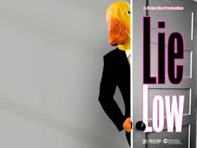 A person in a suit and yellow duck mask stands behind a door. Large pink and white text on the right reads “Lie Low.”.
