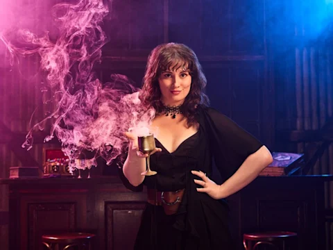 Production photo of Zoe Harlan in a black dress stands at a bar, holding a smoking goblet with pink vapor swirling around it.
