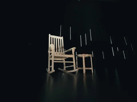 A white wooden rocking chair and a matching small round stool are illuminated in a dark, minimalist room with vertical light streaks on the walls.