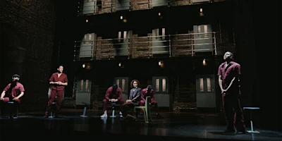 Six people in maroon uniforms are on a dimly lit stage resembling a prison, with two levels of barred doors and balconies in the background.