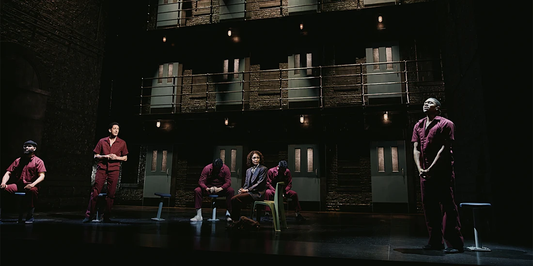 Six people in maroon uniforms are on a dimly lit stage resembling a prison, with two levels of barred doors and balconies in the background.