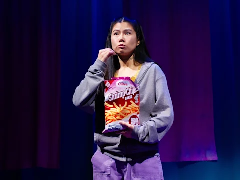 A person stands on stage holding and eating from a bag of shrimp chips.