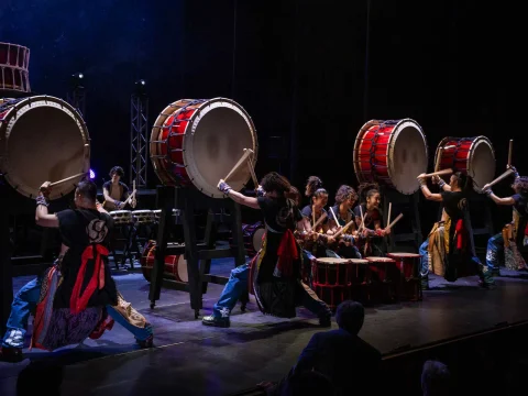 A group of performers in traditional attire playing large taiko drums on a dimly lit stage.