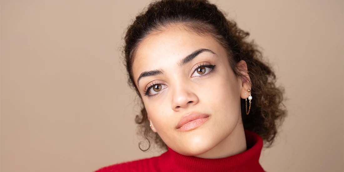 A young woman with curly hair, wearing a red turtleneck and gold hoop earrings, looks at the camera with a neutral expression against a beige background.