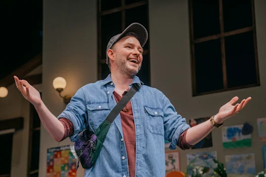 A man in a denim shirt and cap stands indoors with arms open, smiling. Colorful art is displayed on the wall behind him.