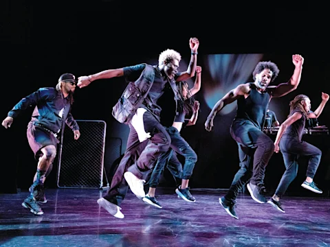 A group of people in dark clothing energetically perform a choreographed dance on stage with a dark backdrop.