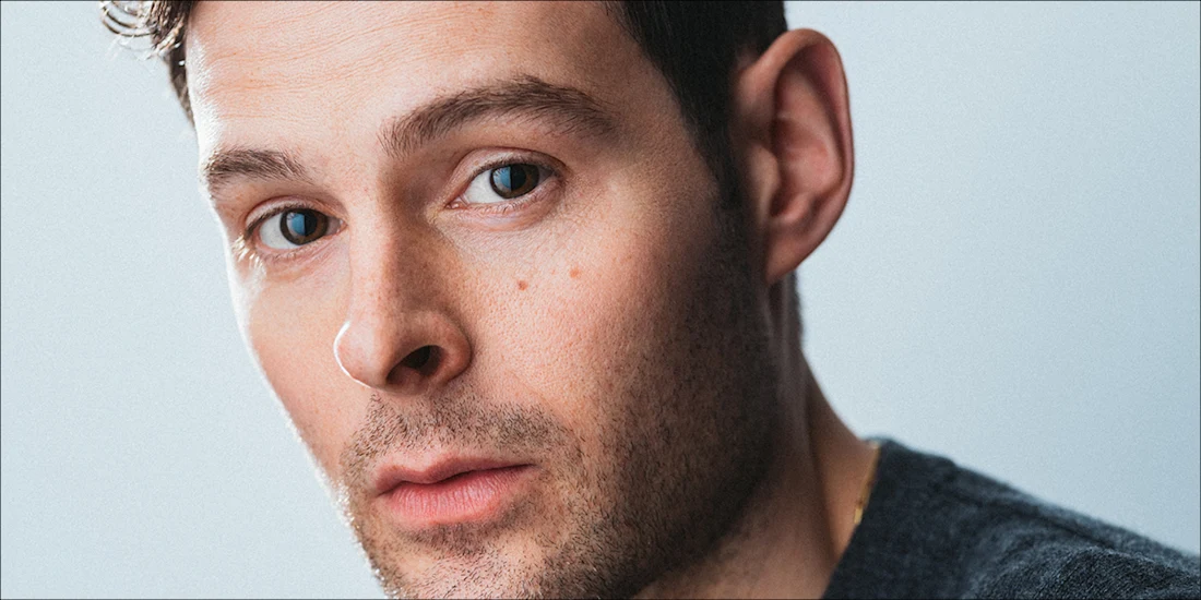 Man with short dark hair, light skin, and blue eyes looks directly at the camera against a plain light background. He is wearing a dark grey sweater.