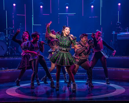 Six performers in colorful, modern costumes sing and dance energetically on a brightly lit stage with musical instruments in the background.