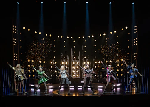 Six performers in colorful, Tudor-inspired costumes sing and gesture on a brightly lit stage with spotlights and confetti in the air.