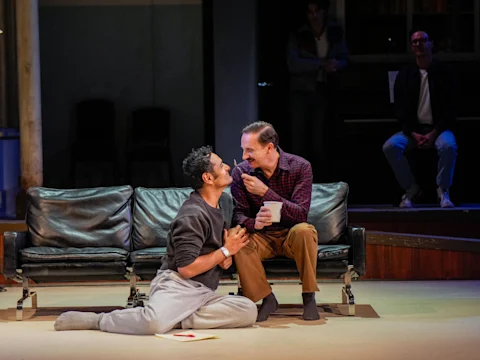 Two men sit closely on the floor in front of a black couch, holding hands and looking at each other; one holds a cup.