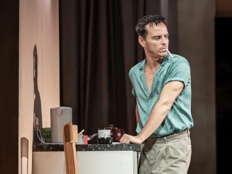 A man in a green shirt stands by a counter, looking to his left with a pensive expression in a well-lit room.