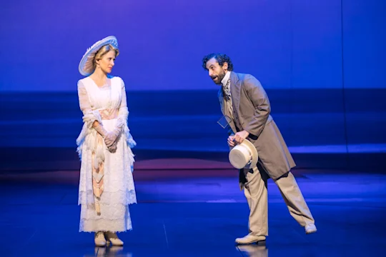 Two actors on stage; the woman wears a white Victorian-era dress and hat, while the man in a suit and scarf bows, holding a straw hat. The background is a blue, minimalist set.