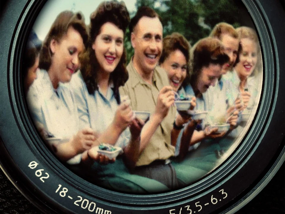 A group of people, mostly women, sit closely together and eat while smiling and laughing. The image is framed to mimic the view through a camera lens with focal length and aperture details.