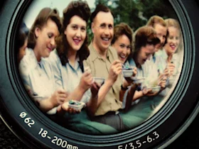 A group of people, mostly women, sit closely together and eat while smiling and laughing. The image is framed to mimic the view through a camera lens with focal length and aperture details.