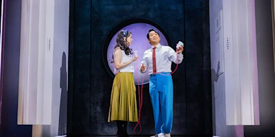 Two actors stand on stage, one holding a microphone and the other holding a prop heart connected by a red cord, with a circular doorway in the background.