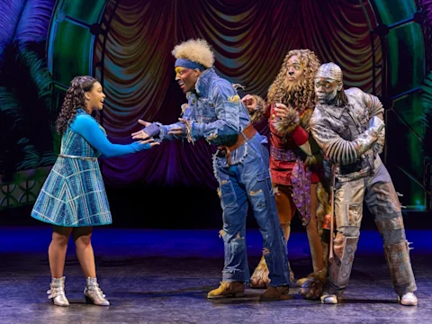 Five actors in colorful costumes perform on stage, with one woman in blue facing four characters dressed as the Scarecrow, Lion, and Tin Man from "The Wizard of Oz.
