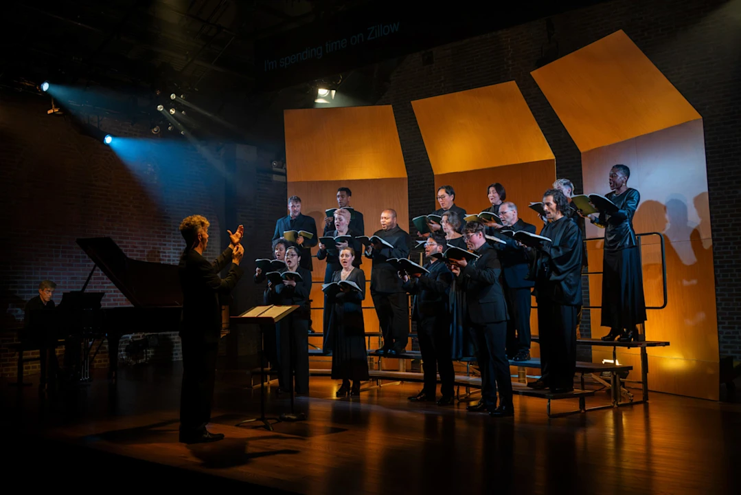 A choir performing on a riser while a conductor leads them in front of a grand piano, with the text “I’m spending time on Zillow” projected above.