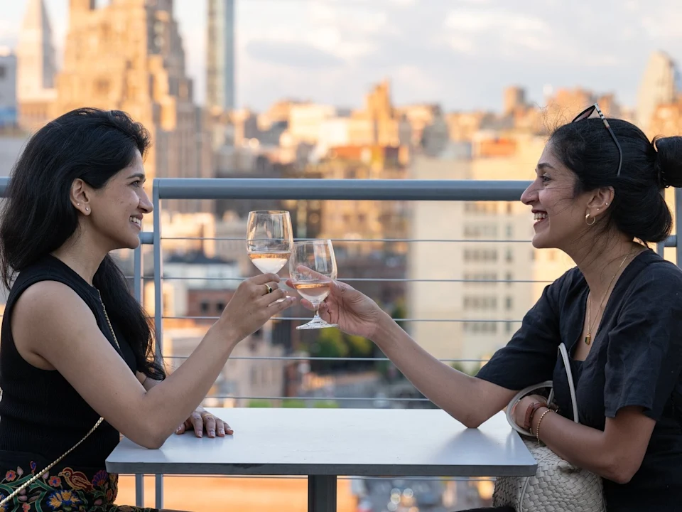 Two women sit at an outdoor table, smiling and clinking wine glasses, with a cityscape in the background.