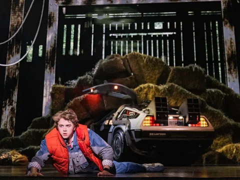 Caden Brauch as Marty McFly in a red vest and denim outfit lies on the ground in front of a DeLorean car with open gull-wing doors, surrounded by a cluttered environment.