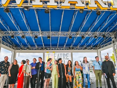 A group of people stand in a row on an outdoor stage under a blue canopy, with a "THEATRE" sign in the background.