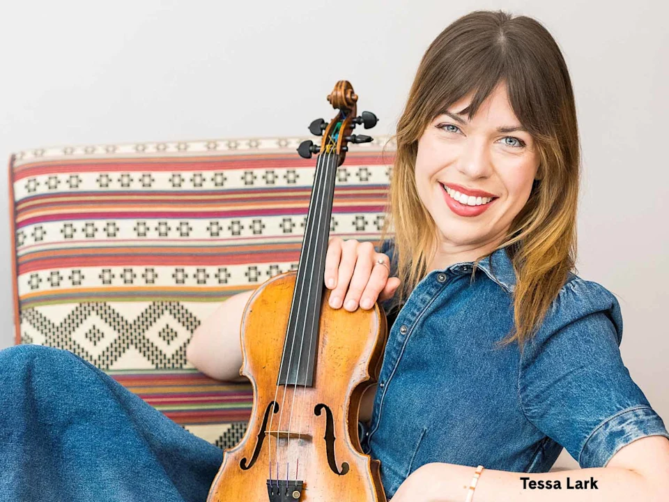 Woman in a blue dress sits on a patterned couch, smiling, and holding a violin with her right hand.