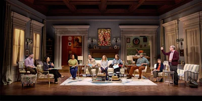 Nine people are seated or standing in a living room set on stage, engaged in conversation, with bookshelves, paintings, and furnishings visible in the background.