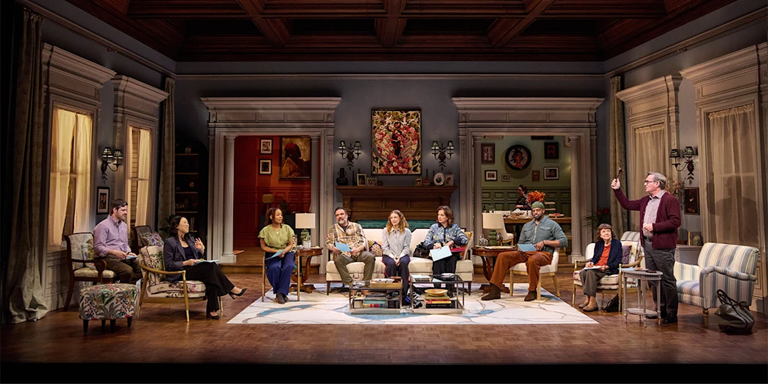 Nine people are seated or standing in a living room set on stage, engaged in conversation, with bookshelves, paintings, and furnishings visible in the background.