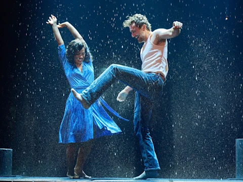 Production photo of The Notebook in Costa Mesa, showing a woman in a blue dress and a man in a white tank top and jeans dance joyfully together in falling water on stage.