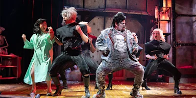 Actors in dramatic costumes perform an energetic dance on stage; one wears a green robe, others are in black, and one in a studded jacket with a skull design stands center.