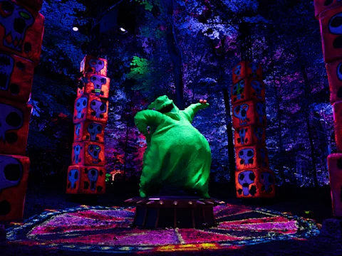 A green, ghostly figure stands on a platform surrounded by glowing totem poles with skull designs. The scene is illuminated by vibrant purple, blue, and pink lights in a wooded area.