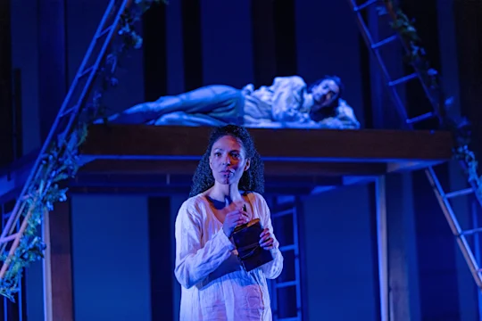Two women in white clothing perform on a stage; one stands holding a book in the foreground, while the other lies on an elevated platform in the background under blue lighting.