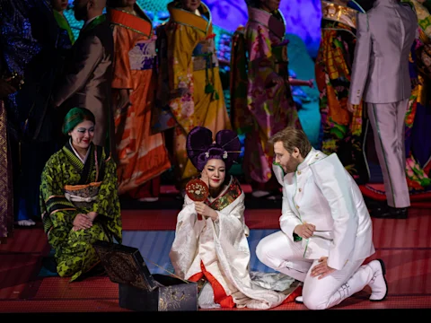 A woman in elaborate white and purple attire holds a mirror, surrounded by people in colorful kimonos and white suits on stage.