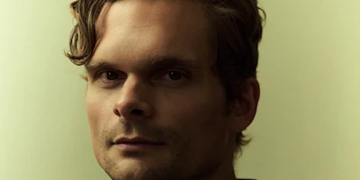 A man with wavy hair looks directly at the camera against a plain, softly lit background.