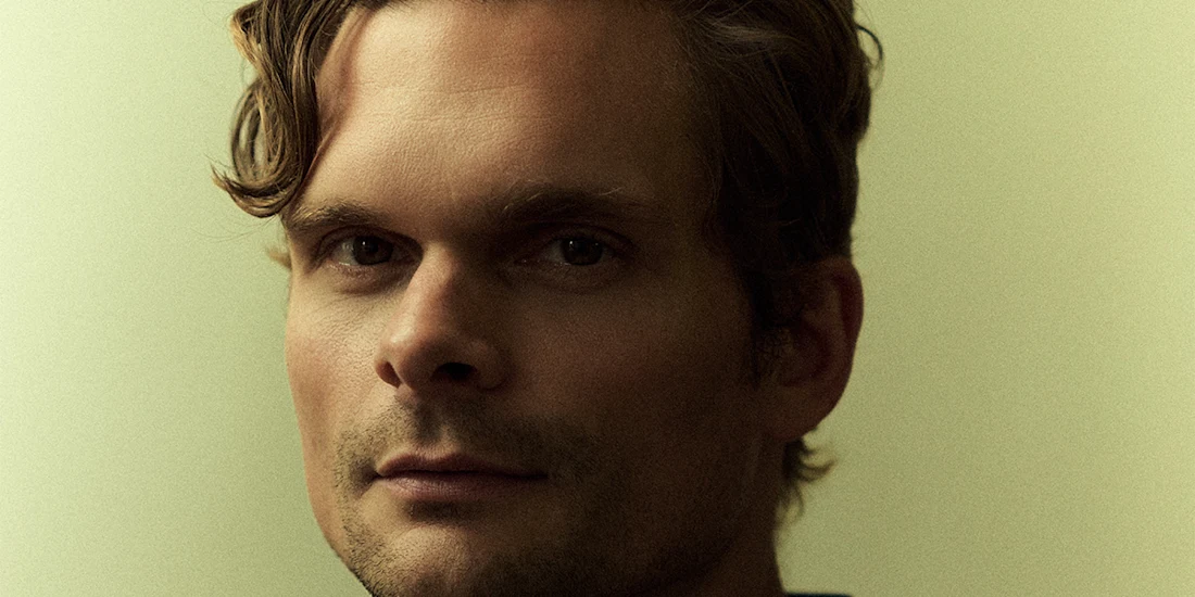 A man with wavy hair looks directly at the camera against a plain, softly lit background.