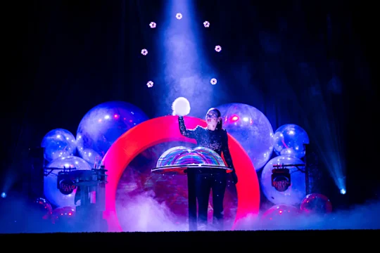 A performer on stage stands in front of large, illuminated bubbles, holding a glowing sphere with one hand under dramatic lighting and colorful effects.