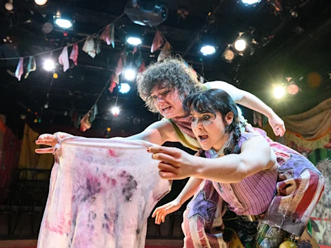 Two actors on stage, one leaning over the other, both with intense expressions, holding a stained cloth under bright stage lights with festive decorations in the background.