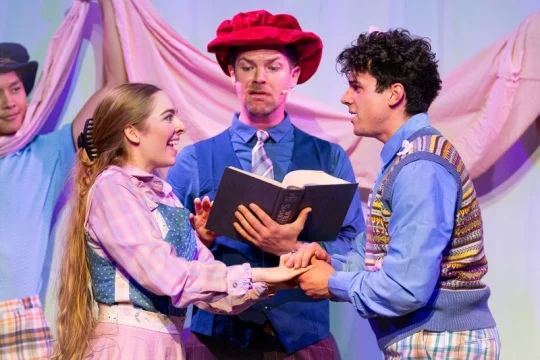 A man and woman in colorful costumes hold hands and smile while another man in a hat reads from a book, all on stage during a theatrical performance.