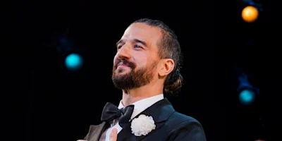 A man with a beard in formal attire, wearing a black tuxedo, bow tie, and white flower, stands against a dark background with colorful lights.