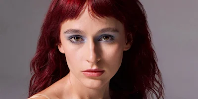 Person with long red hair and blue eyeshadow looks directly at the camera against a neutral background.