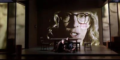 Two people sit on the floor of a stage in front of a table, with a large, ghostly face projected onto the wall behind them.