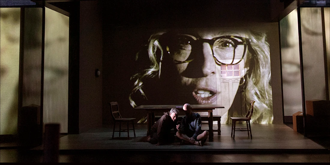 Two people sit on the floor of a stage in front of a table, with a large, ghostly face projected onto the wall behind them.